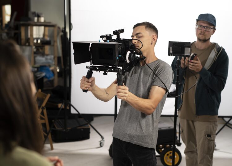 Foto seorang pria sedang memegang kamera profesional dalam set film, dikelilingi alat-alat serta kru film lainnya.